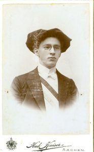 Glattrasierter junger Mann, mit Anzug, rot-weiss-blauer Schärpe, Barrett und Brille.